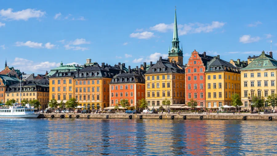 Die ockerfarbenen, orange und roten Kaufmannshäuser der Altstadt Gamla Stan vom Wasser aus entlang Skeppsbron, ein Kirchturm im Hintergrund.