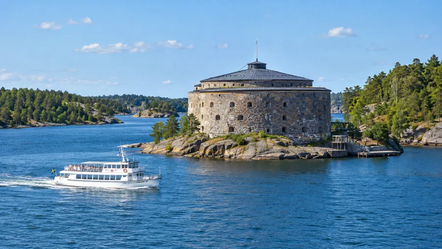 Eine runde Steinfestung auf einer kleinen Felseninsel in einer engen Meerenge, ein weißes Passagierboot passiert sie